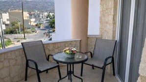 A balcony with two chairs and a table, a flower vase, and a view of a street and buildings. - Sun Studio - Sitia Cozy Stay With Mountain Views (Sitia)