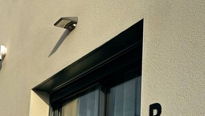 Eine Gebäudefassade mit einem großen Fenster, einem Schild mit dem schwarzen Buchstaben 'B' und einer Überwachungskamera.