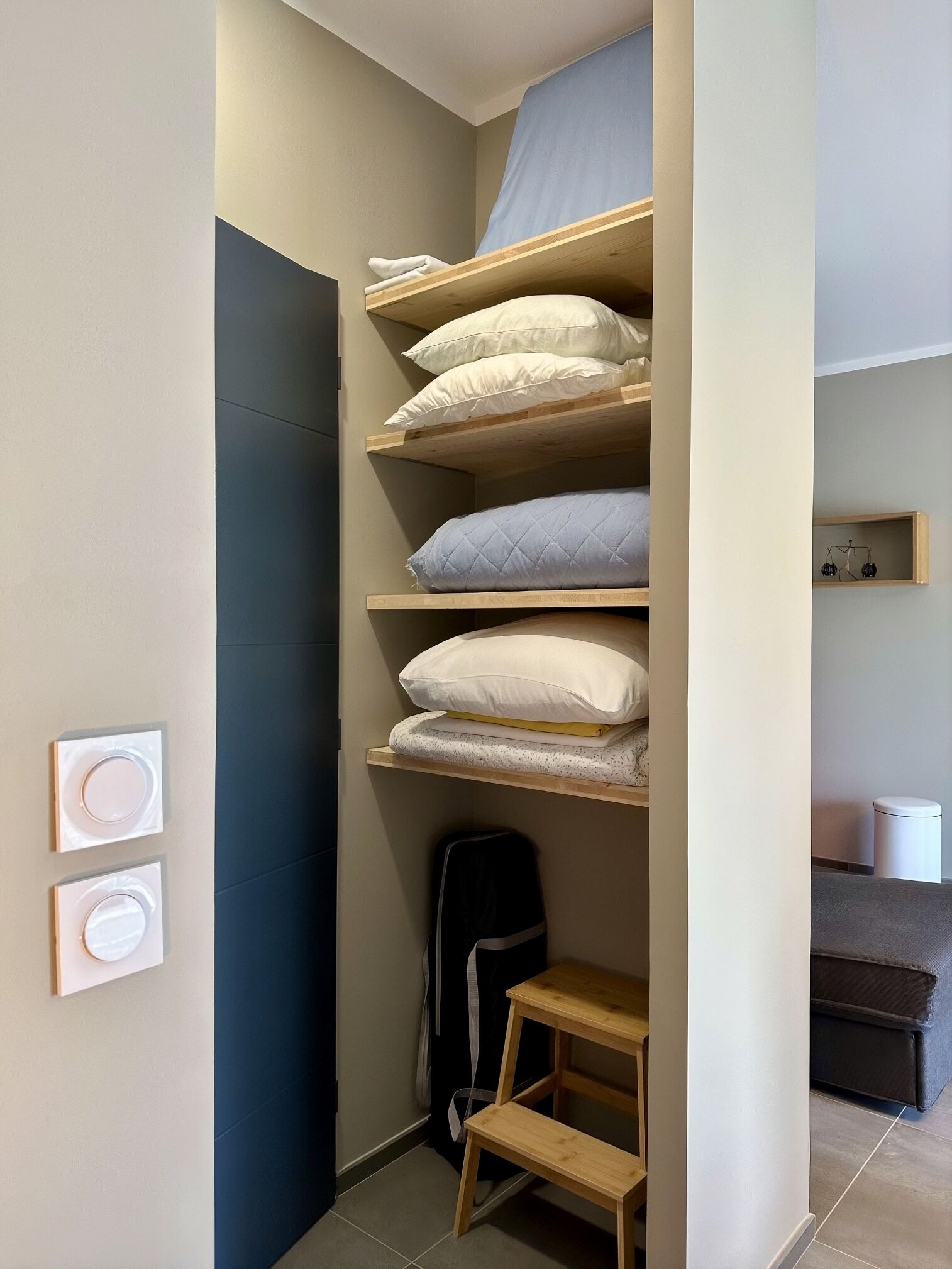Een kleine kamer met een stapelbed, een klein houten krukje en een wandplank met decoratieve spullen.