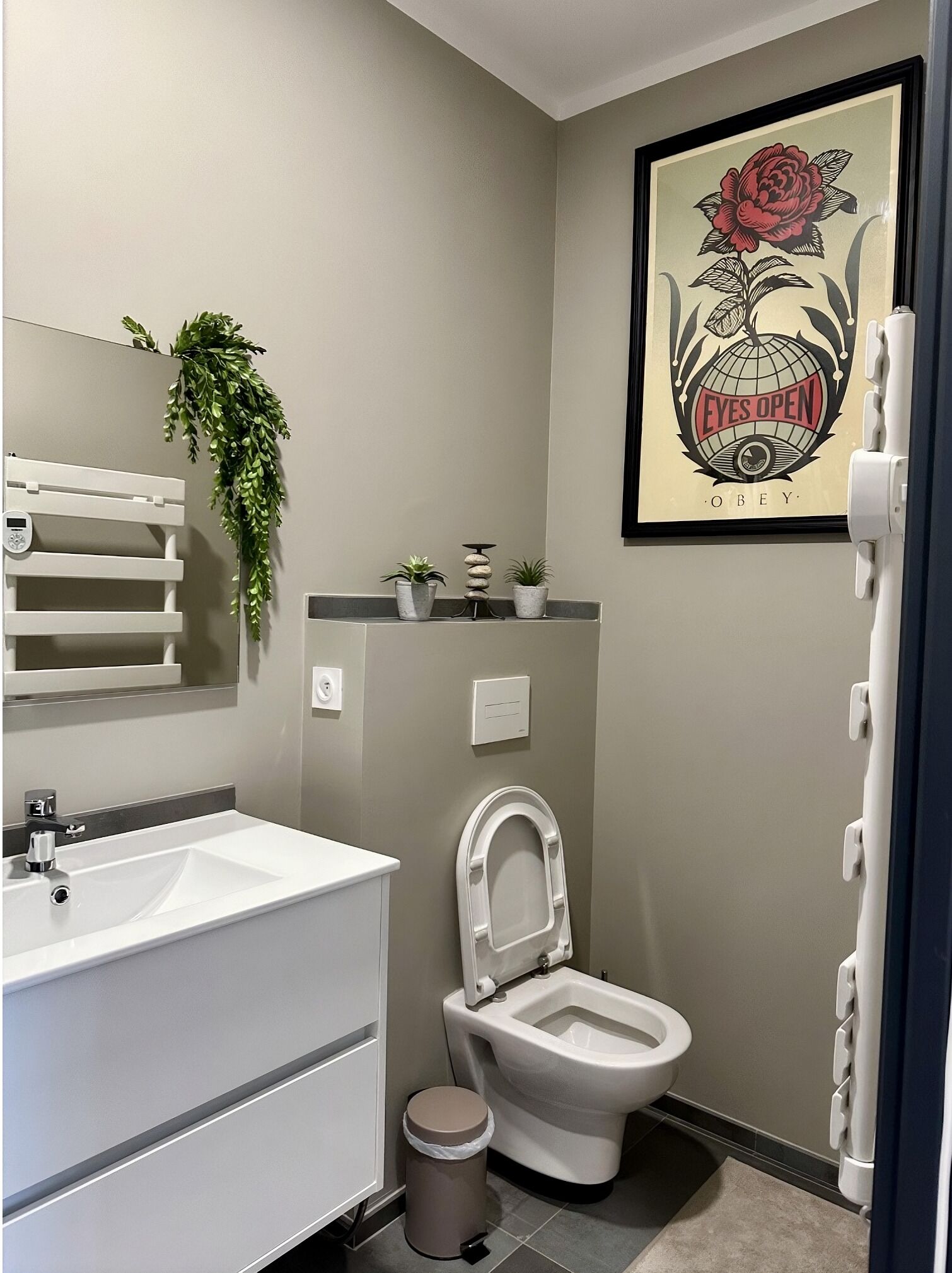 Un baño moderno con un lavamanos blanco, un inodoro, una obra de arte enmarcada en la pared y una pequeña planta en maceta sobre el mostrador.