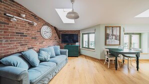 Ein gemütliches Wohnzimmer mit Backsteinwand, einem grünen Sofa, einem Esstisch mit Stühlen, einem Fernseher und einem Oberlicht.