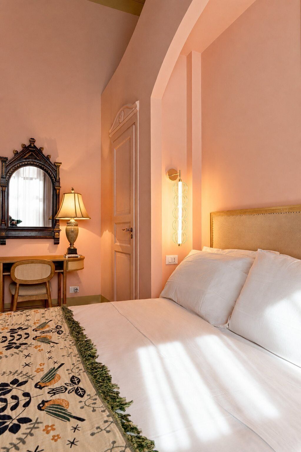 A hotel room with a bed, a desk with a lamp, a mirror, and a wall-mounted light fixture.