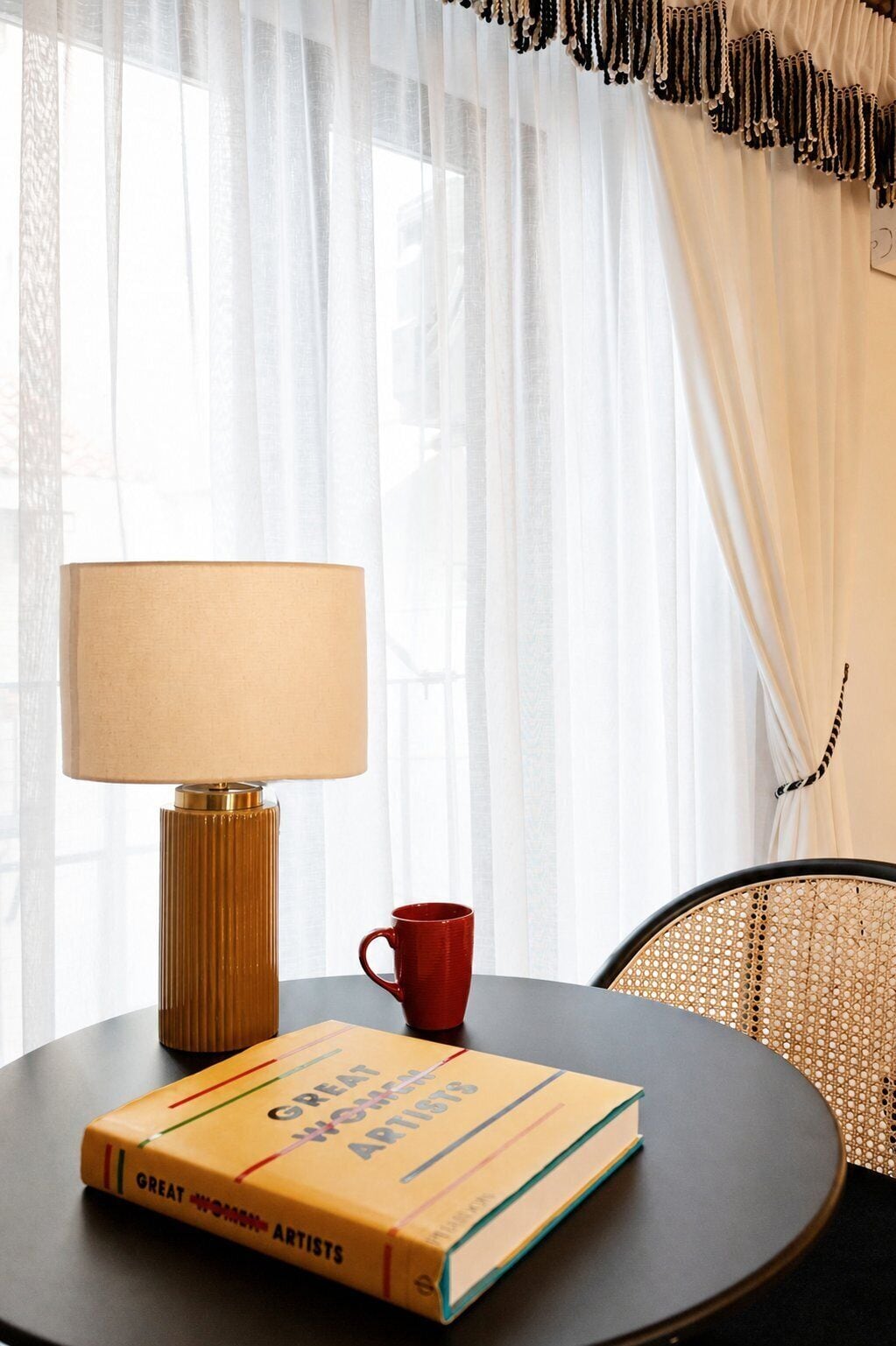 Una mesa redonda con una lámpara, una taza roja y un libro titulado 'Great Women Artists' en una habitación con cortinas traslúcidas y una silla estampada.
