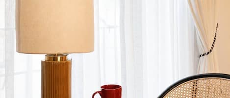 A round table with a lamp, a red mug, and a book titled 'Great Women Artists' in a room with sheer curtains and a patterned chair.