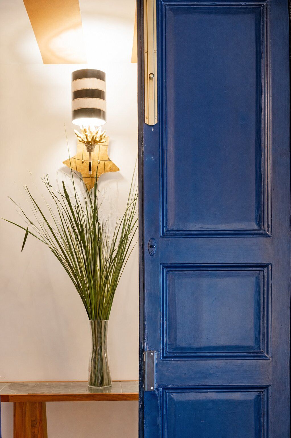 A blue door, a vase with green plants, and a wall-mounted lamp.