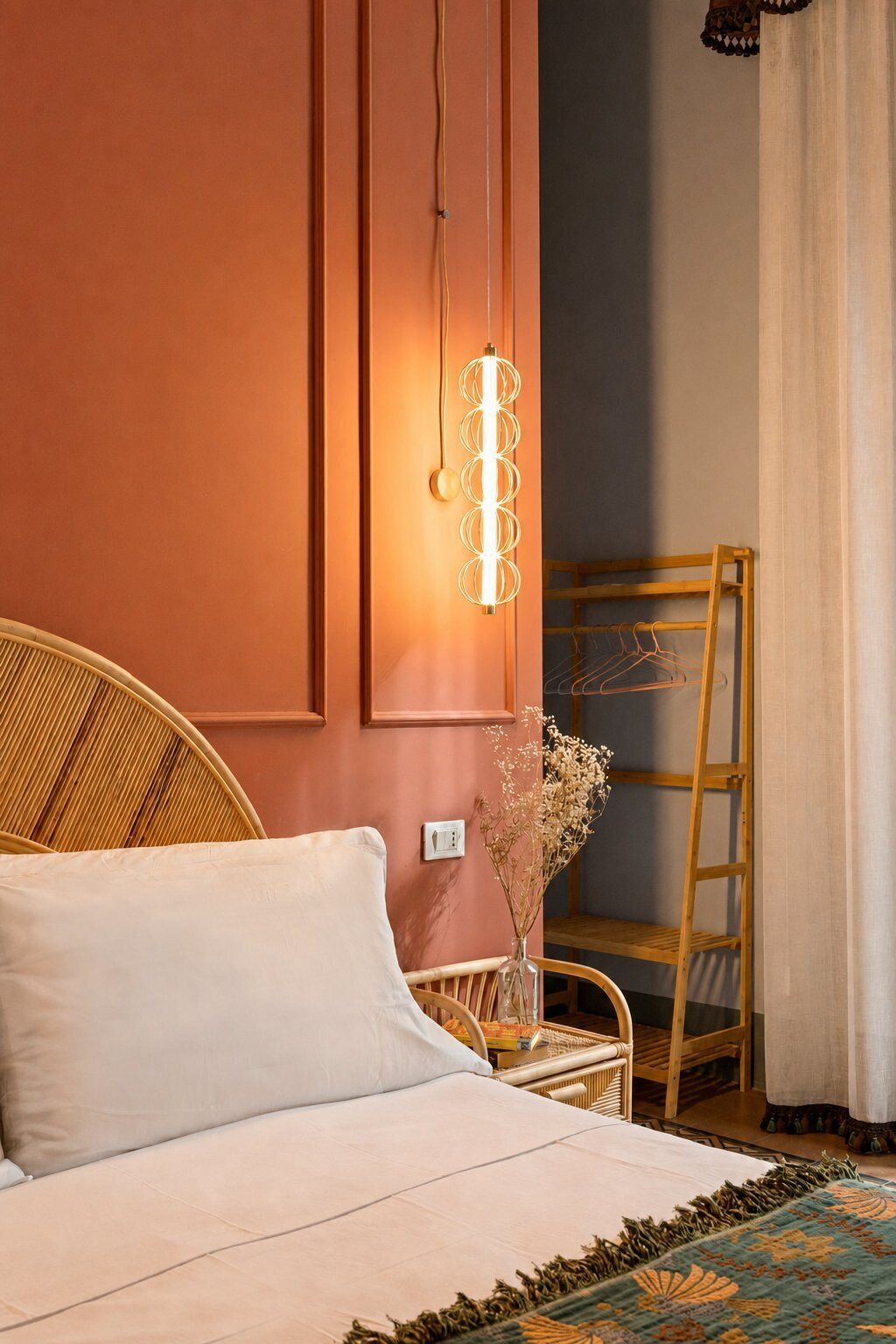 A bedroom with a bed, a wicker chair, a wooden ladder, a wall-mounted light fixture, and a vase with dried flowers.
