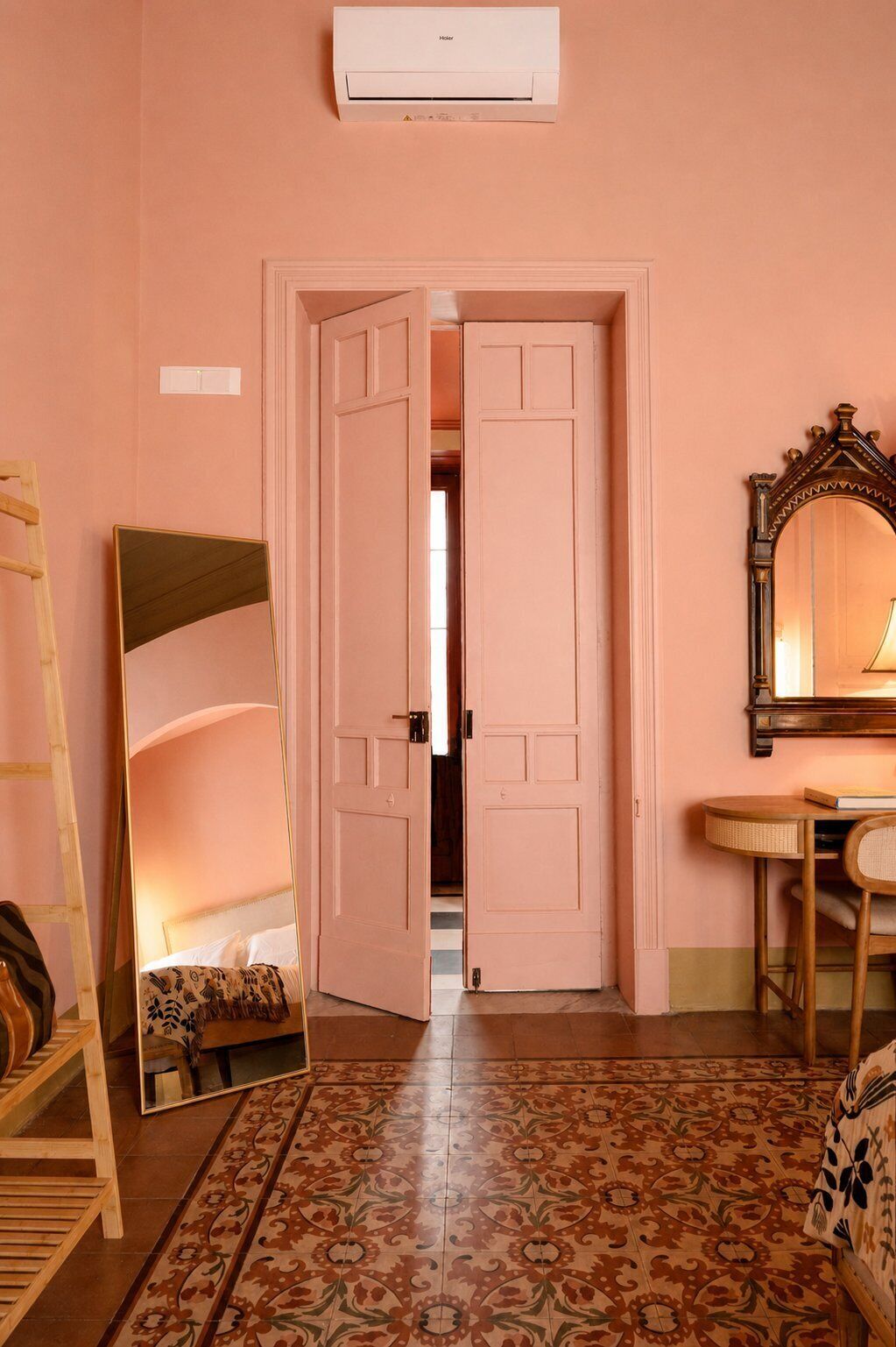 Una habitación con paredes rosas, un espejo grande, una escalera de madera, una alfombra estampada y una cama con cabecera.