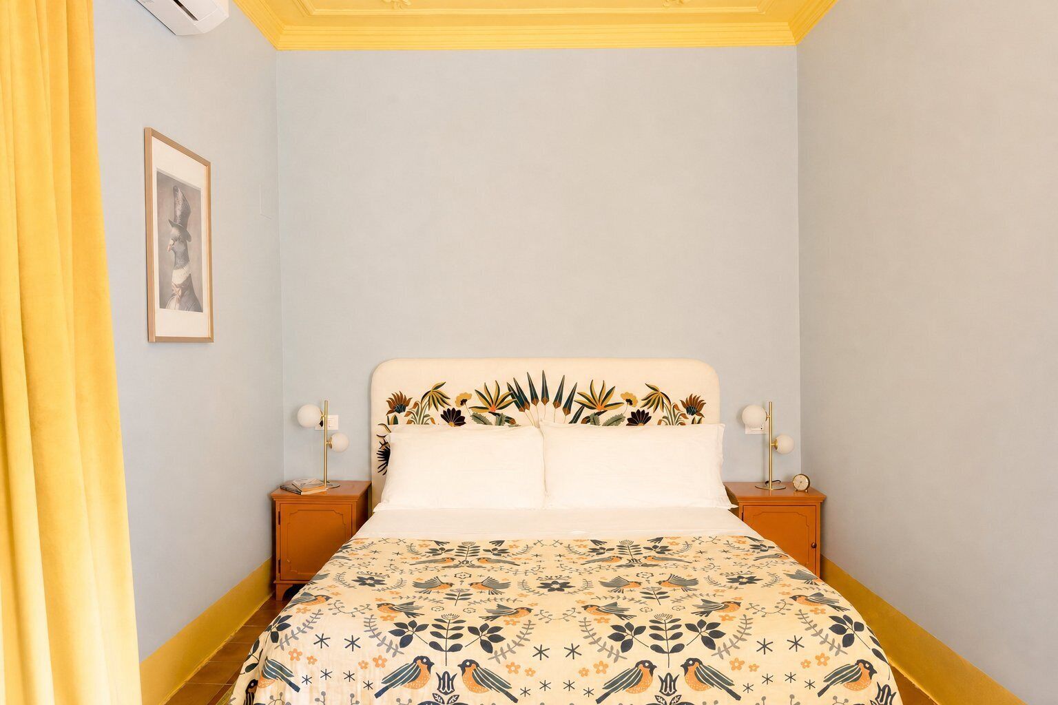 A bedroom with a bed, two bedside tables, a nightstand, a framed picture on the wall, and a window with curtains.