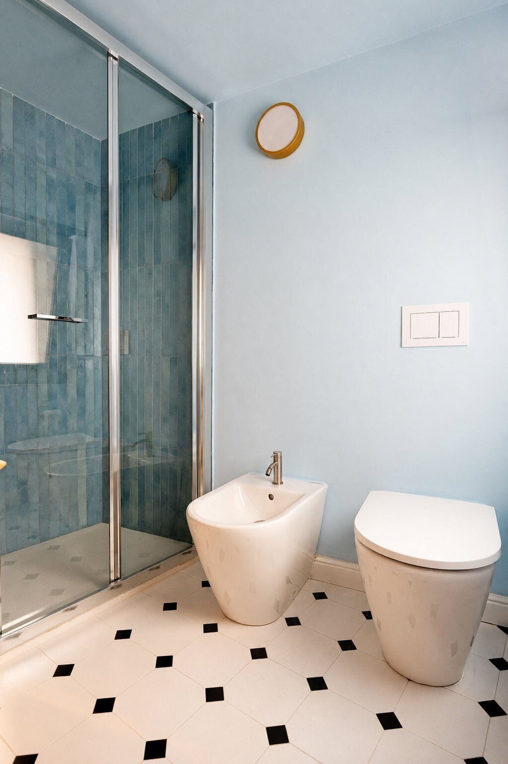 A bathroom with a glass shower enclosure, a bidet, a toilet, and a light switch on the wall.