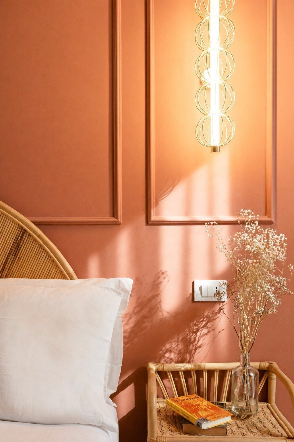 Een kamer met een bed, een wandlamp, een nachtkastje met een boek en een vaas gedroogde bloemen, en een lichtschakelaar.