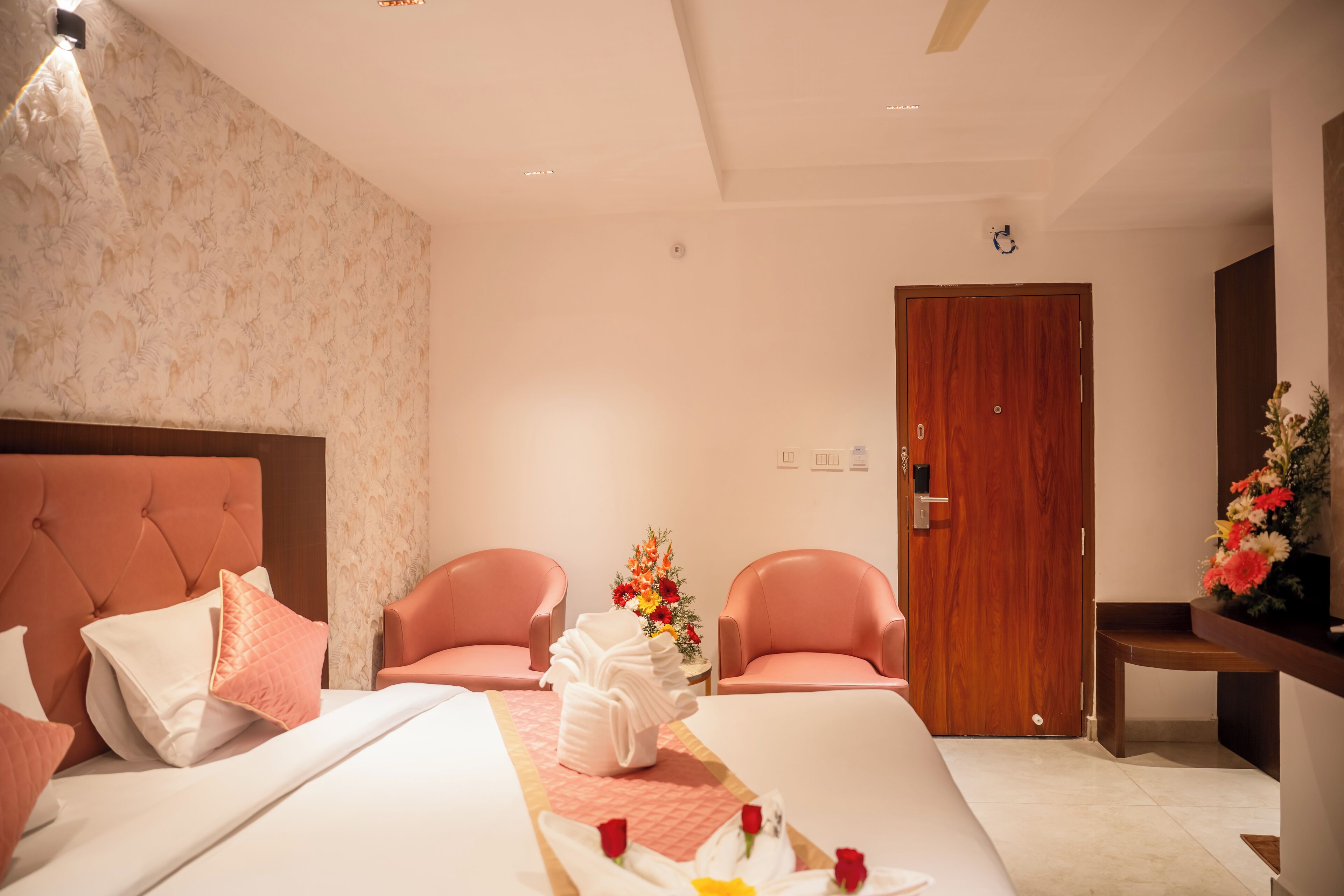 Habitación de hotel con una cama grande, dos sillones rosados, una puerta de madera y una pequeña mesa con un jarrón de flores.
