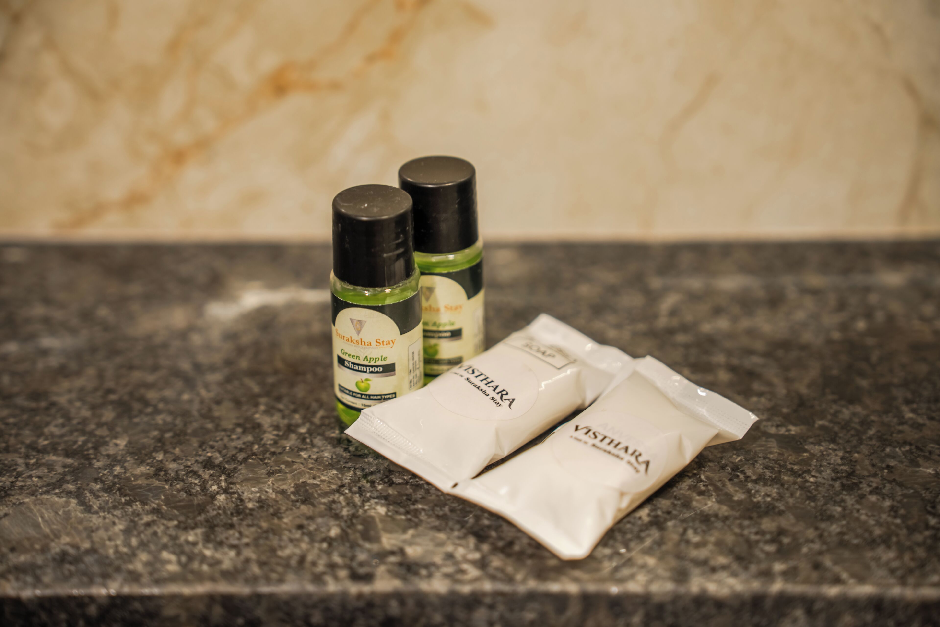Two bottles of green apple shampoo and two sachets of hair care product on a granite countertop.