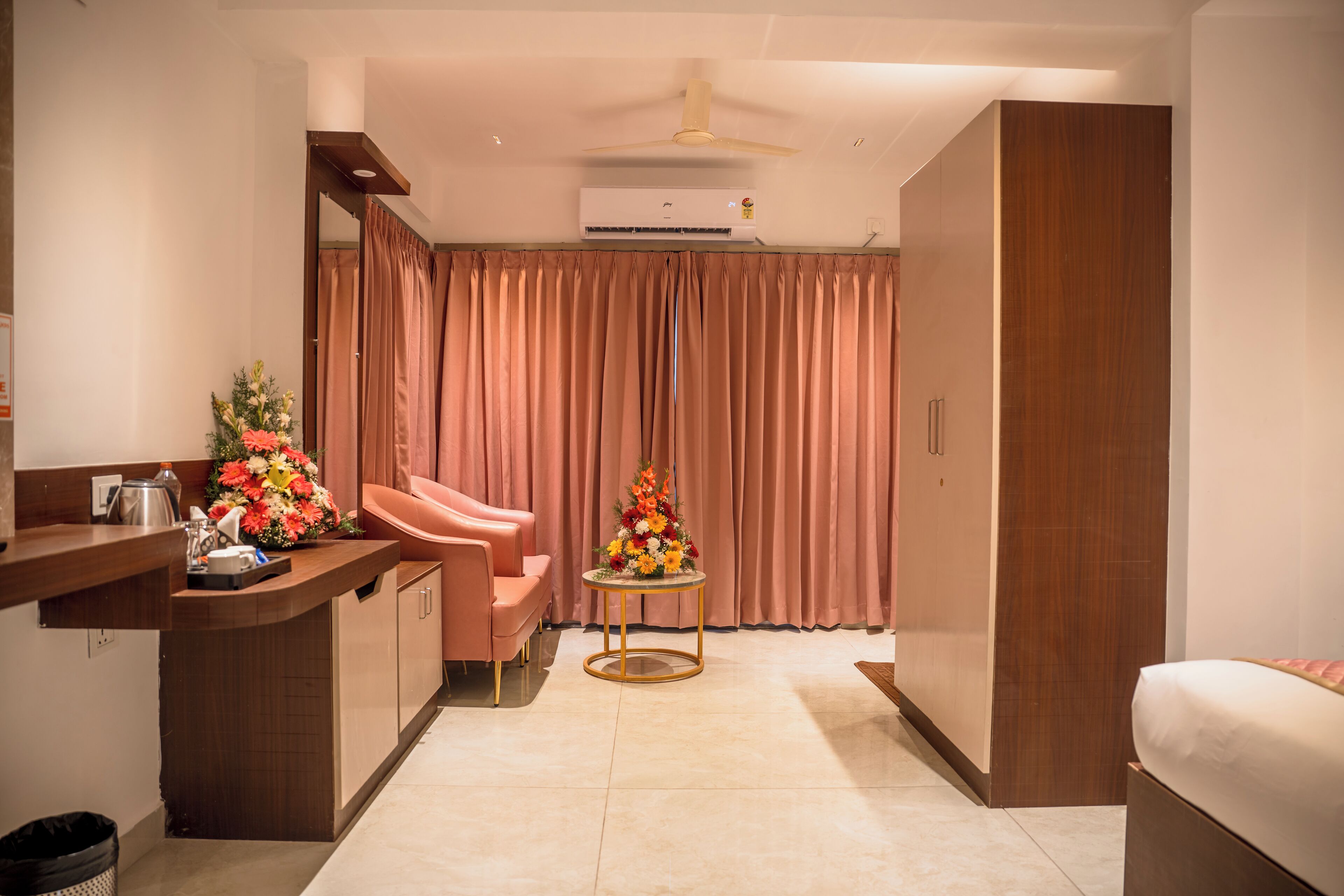 Une chambre d’hôtel équipée d’une kitchenette, d’un lit, d’un coin salon avec un fauteuil rose, d’une petite table ronde agrémentée d’un centre de table floral et d’une grande fenêtre avec des rideaux.