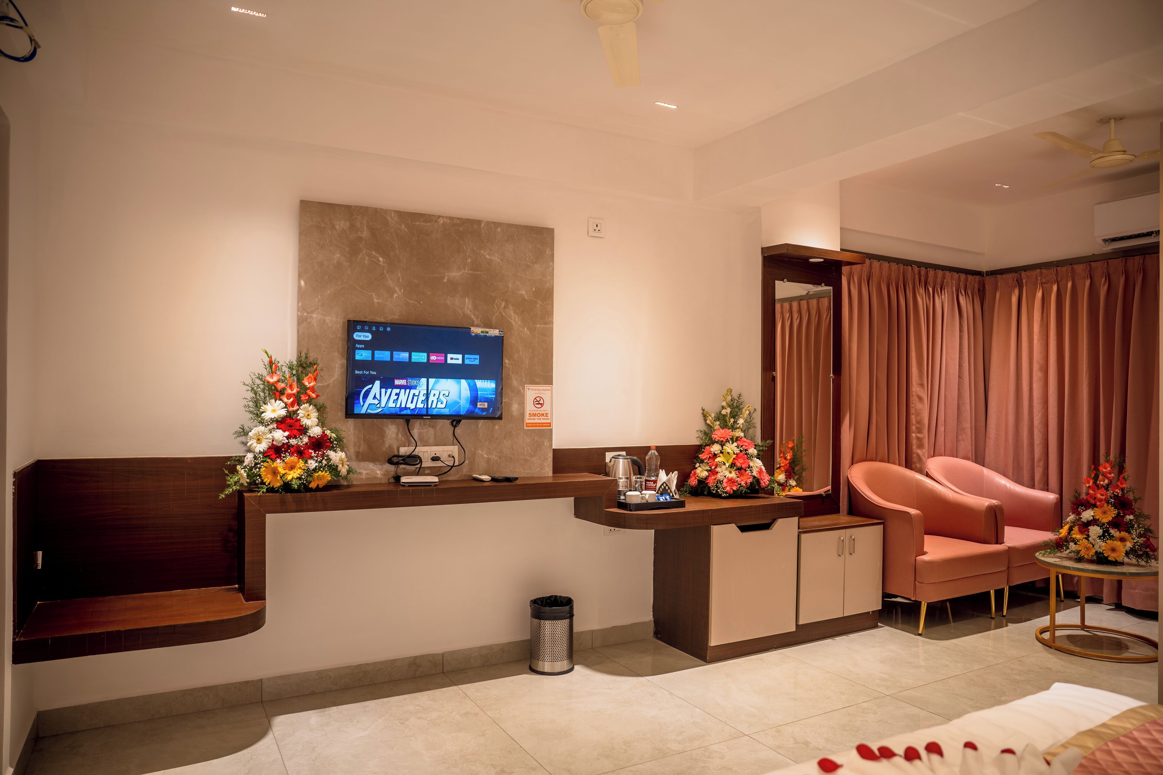 A modern hotel lobby with a reception desk, a flat-screen TV displaying 'Avengers', a trash bin, a sofa, and a small table with a vase of flowers.