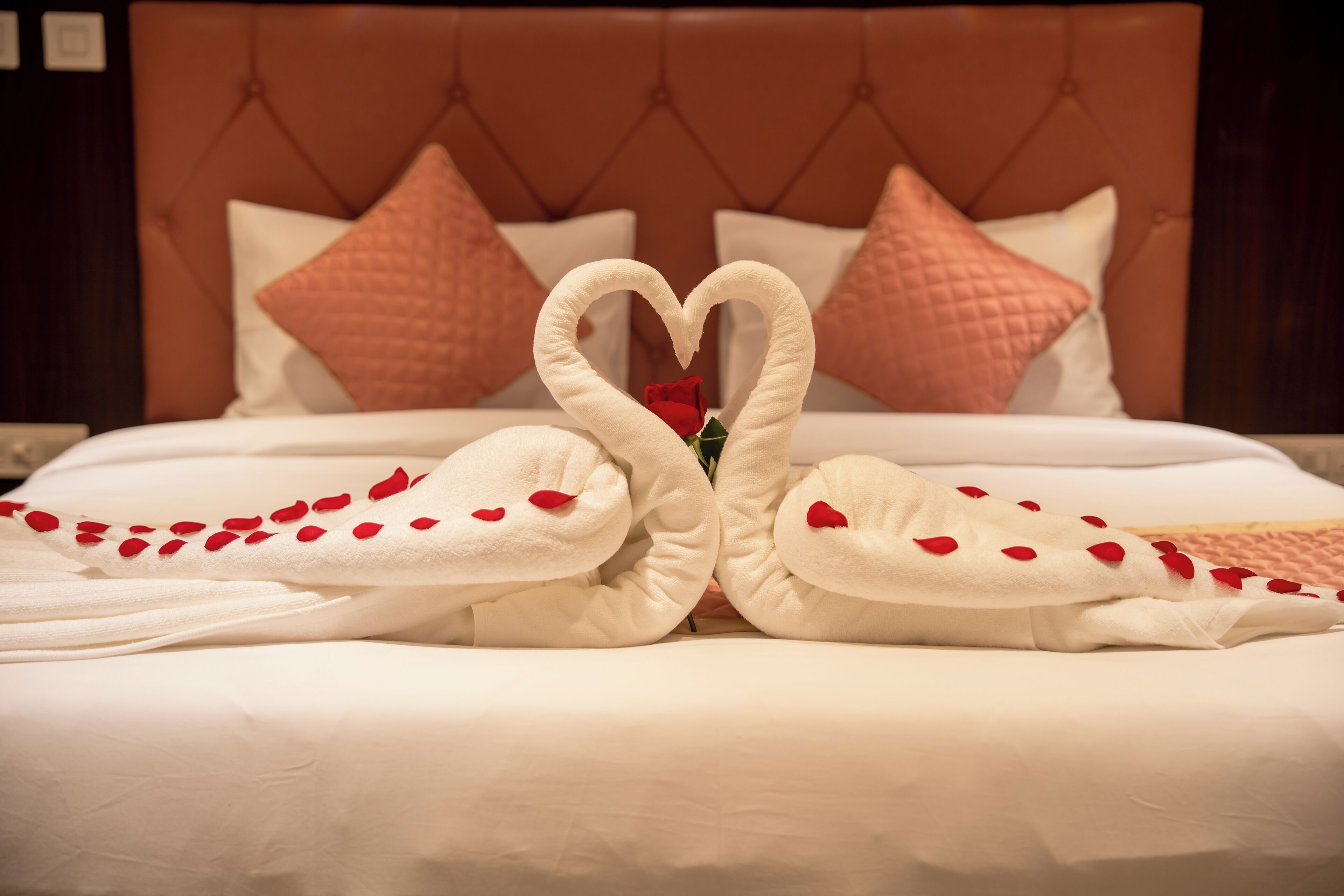 A bed with a heart-shaped towel arrangement and rose petals.