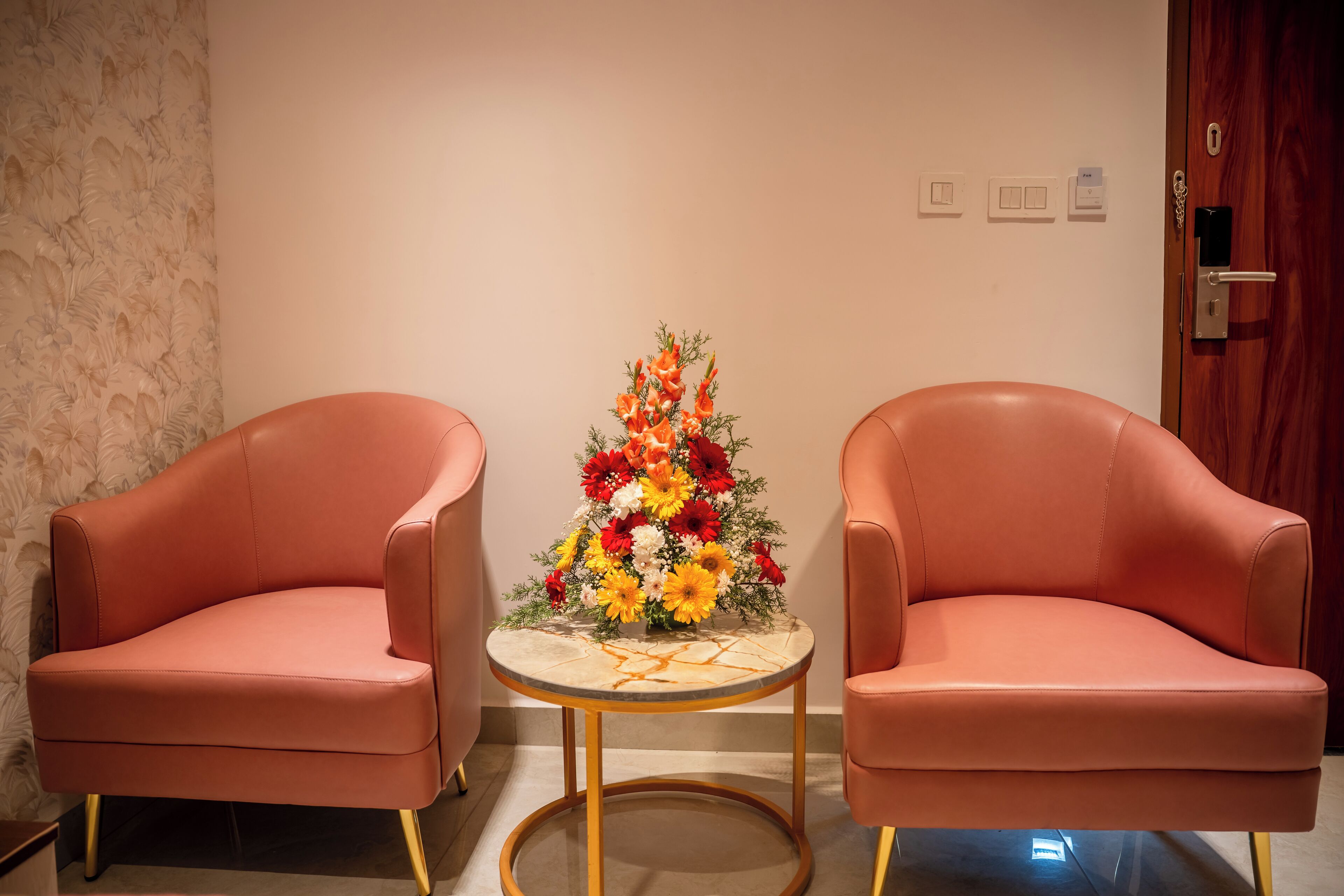 Una habitación con dos sillones de cuero rosa, una mesita redonda con un centro de mesa floral y una puerta de madera con cerradura de llave.
