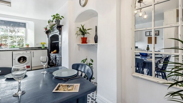 Un coin repas avec une table dressée pour deux personnes, un sol à motifs noir et blanc, un mur blanc orné d’une horloge et une fenêtre donnant sur l’extérieur.