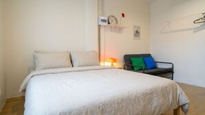 A bedroom with a bed, a bedside table with a lamp, a chair with cushions, a wall-mounted clock, and a picture.