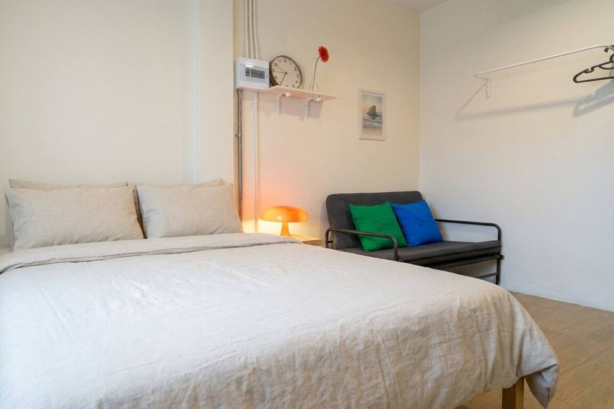 Un dormitorio con una cama, una mesita de noche con lámpara, una silla con cojines, un reloj en la pared y un cuadro.