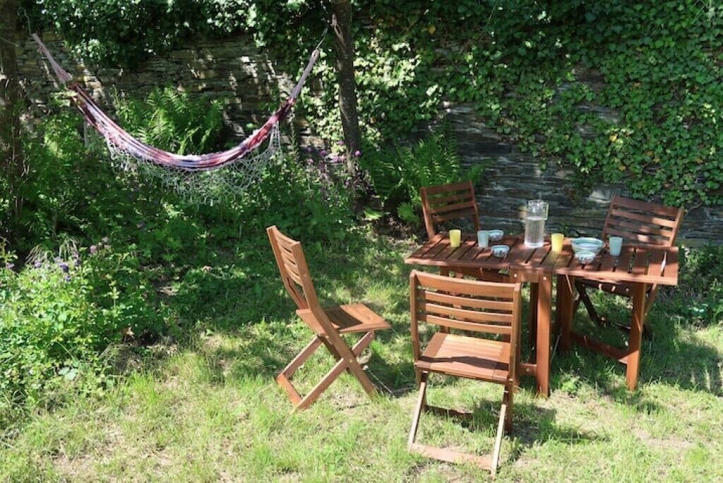 An outdoor wooden table with chairs, a hammock, and a stone wall in the background.