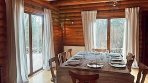 Comedor de cabaña de madera con una mesa dispuesta para seis comensales, ventanas amplias y una lámpara colgante.
