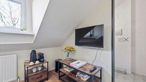Una habitación pequeña y moderna con techo inclinado, un televisor montado en la pared, una mesa de centro de madera con libros y un florero, un radiador y una ventana con vistas a árboles desnudos.