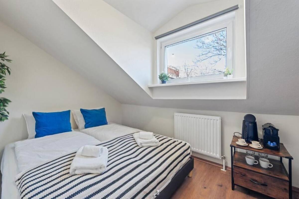 A bedroom with a sloped ceiling, a bed with striped bedding, blue pillows, a radiator, a small table with cups and a coffee maker, and a window with a view of trees and buildings.