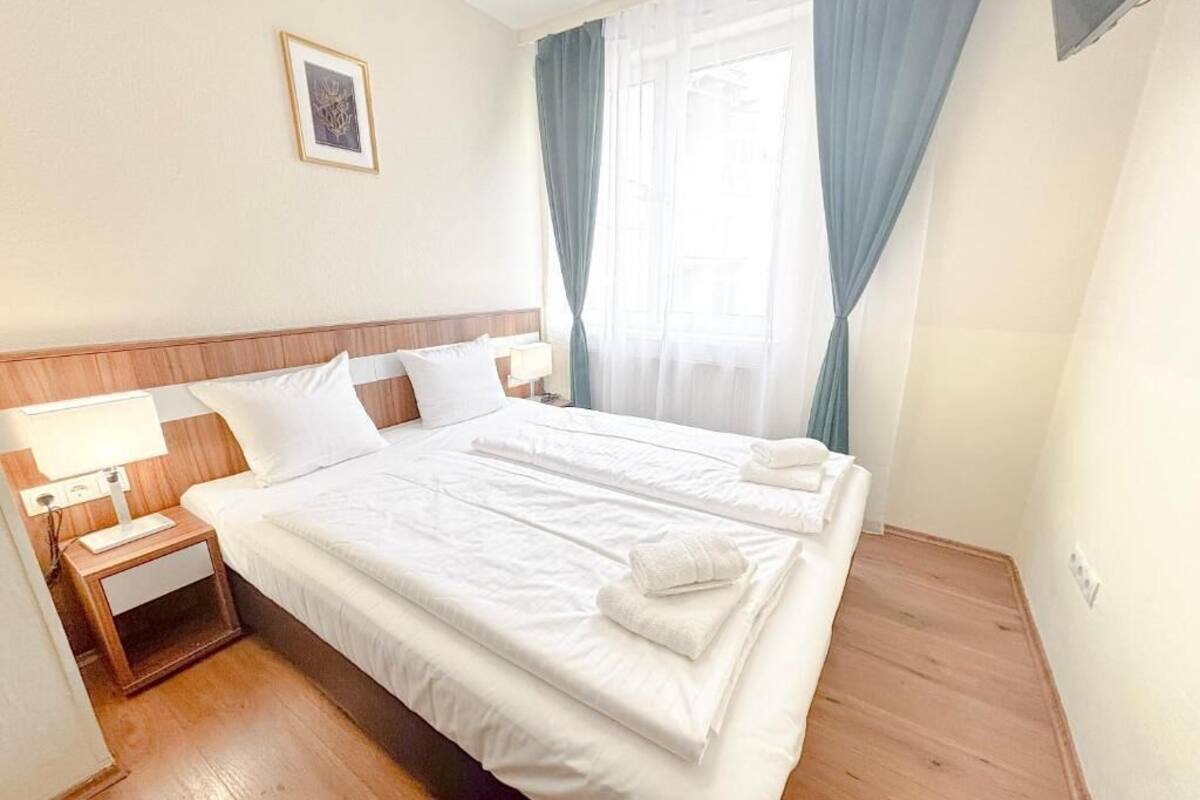 A neatly made bed with white linens and towels, a bedside table with a lamp, and a window with curtains.