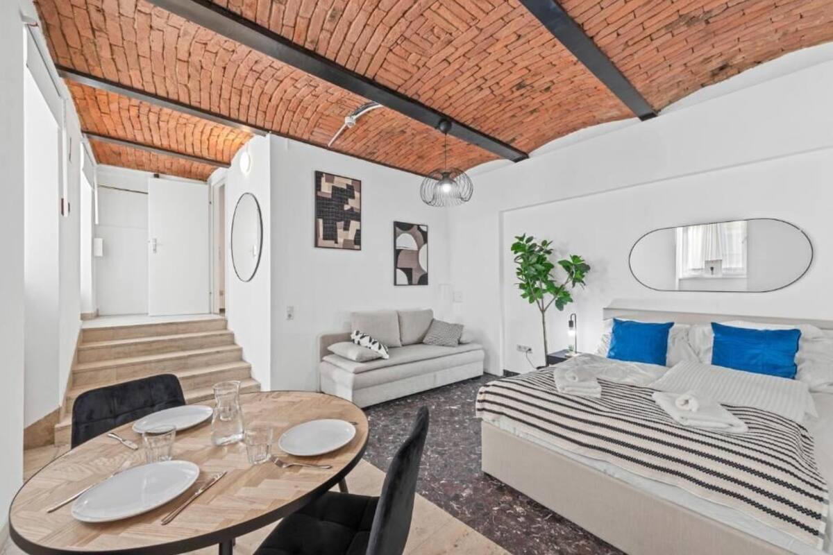 A modern, well-lit room with a brick ceiling, a dining area with a round table and chairs, a sofa, a bed with striped bedding, and a large mirror.