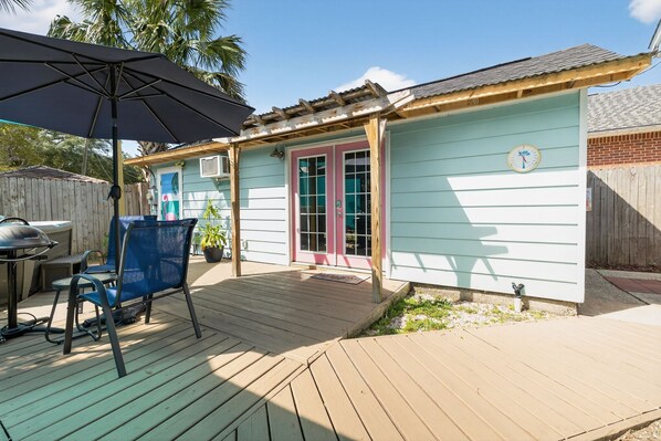 Un jardin avec une terrasse en bois, un barbecue, une chaise bleue et un parasol.