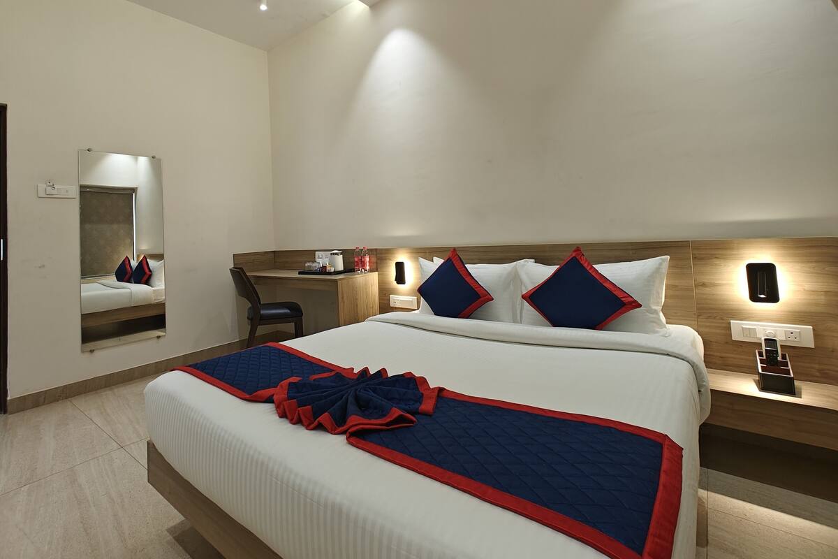 A hotel room with a large bed, a desk, a chair, a mirror, and a nightstand with a phone.