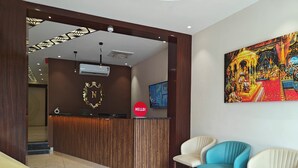 A reception area with a wooden reception desk, a "HELLO!" sign, a mounted television, and a colorful painting on the wall. - Hotel Shivam by Greenwoods Resorts (Kudal)