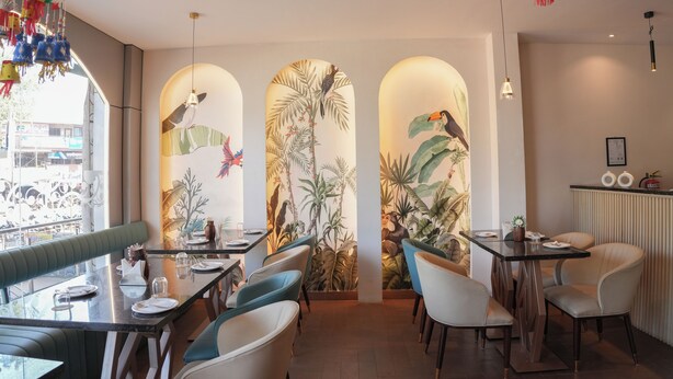 A restaurant with a tropical mural, green seating, and a view of the street. - Hotel Shivam by Greenwoods Resorts (Kudal)
