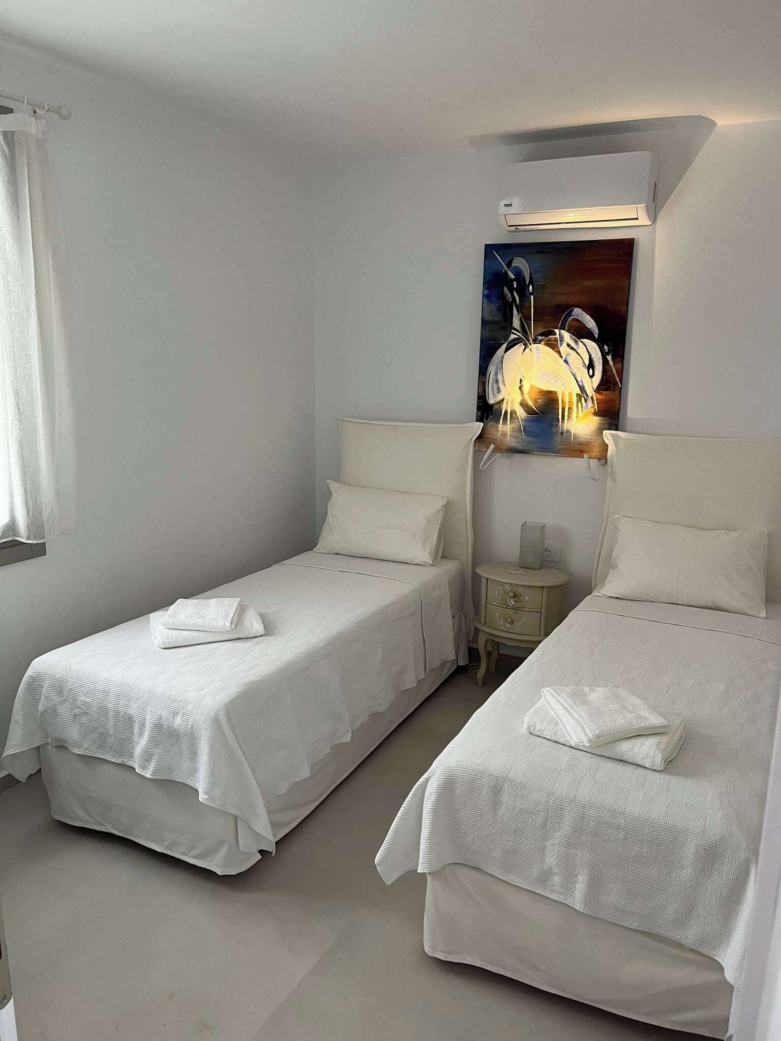 A hotel room with two single beds, each with a white bedspread and pillows, a bedside table with a lamp, and a painting above the headboard.