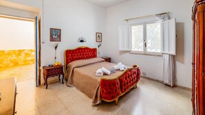 A room with a red tufted bed, a small bedside table, a window with curtains, and a door leading to another room. - Villino Salentum With Private Pool and Garden (Racale)