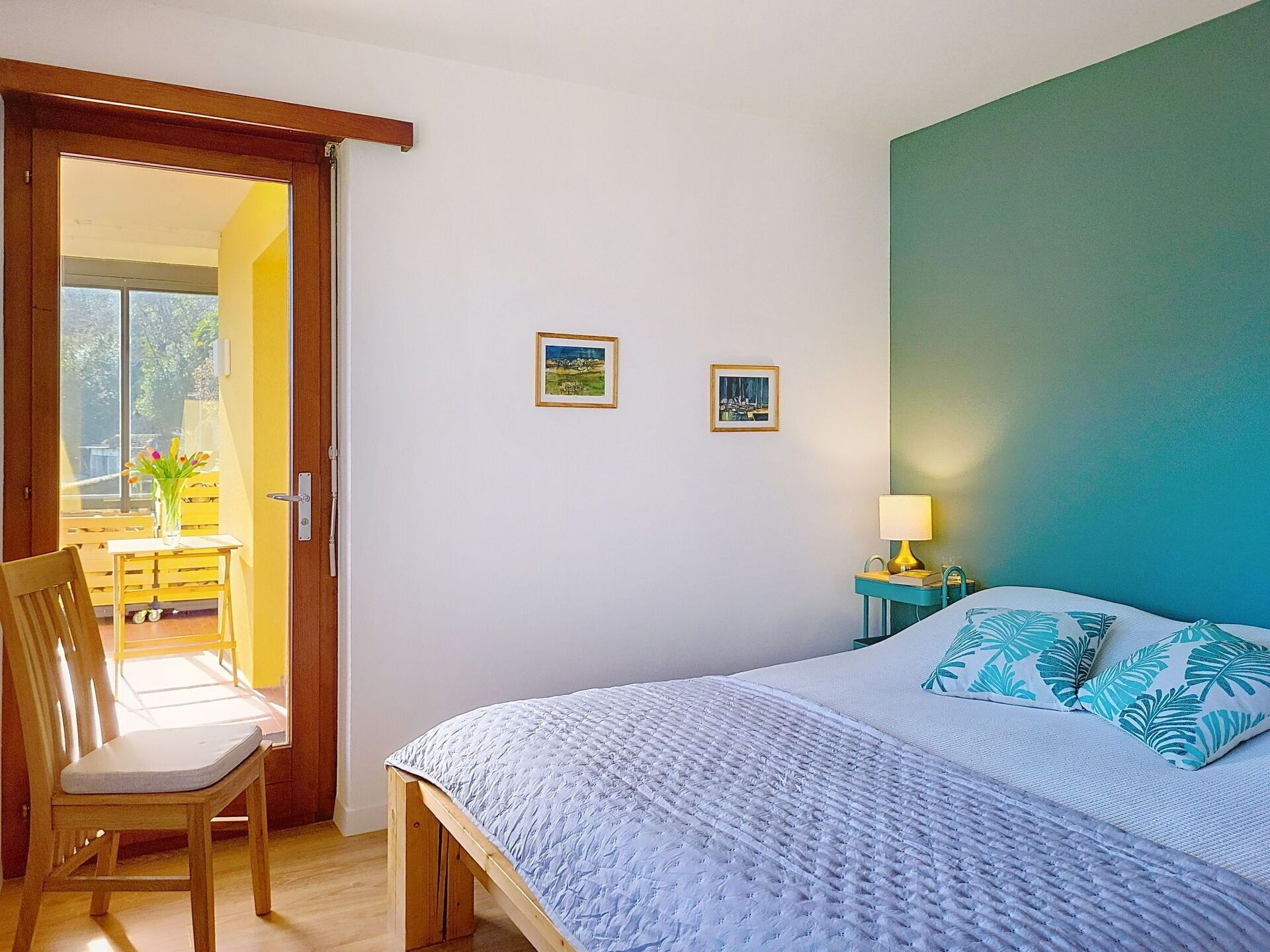 Un dormitorio con cama, una silla, una mesa con un florero y dos cuadros en la pared.