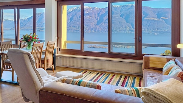 Ein Zimmer mit großem Fenster und Blick auf Berge und See, einem Essbereich mit Holzkästen, einer Couch mit gestreiften Kissen sowie einem Heizkörper unter dem Fenster.