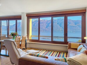 Ein Zimmer mit großem Fenster und Blick auf Berge und See, einem Essbereich mit Holzkästen, einer Couch mit gestreiften Kissen sowie einem Heizkörper unter dem Fenster.