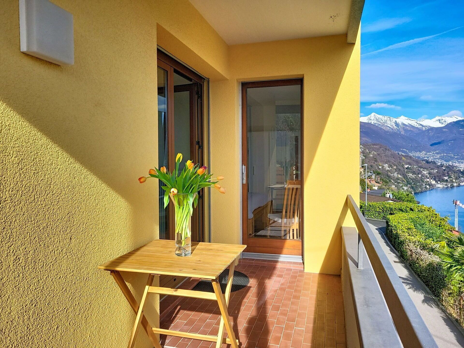 Un balcón con una mesa de madera y un florero, una puerta corredera de vidrio y vistas a un lago y montañas.