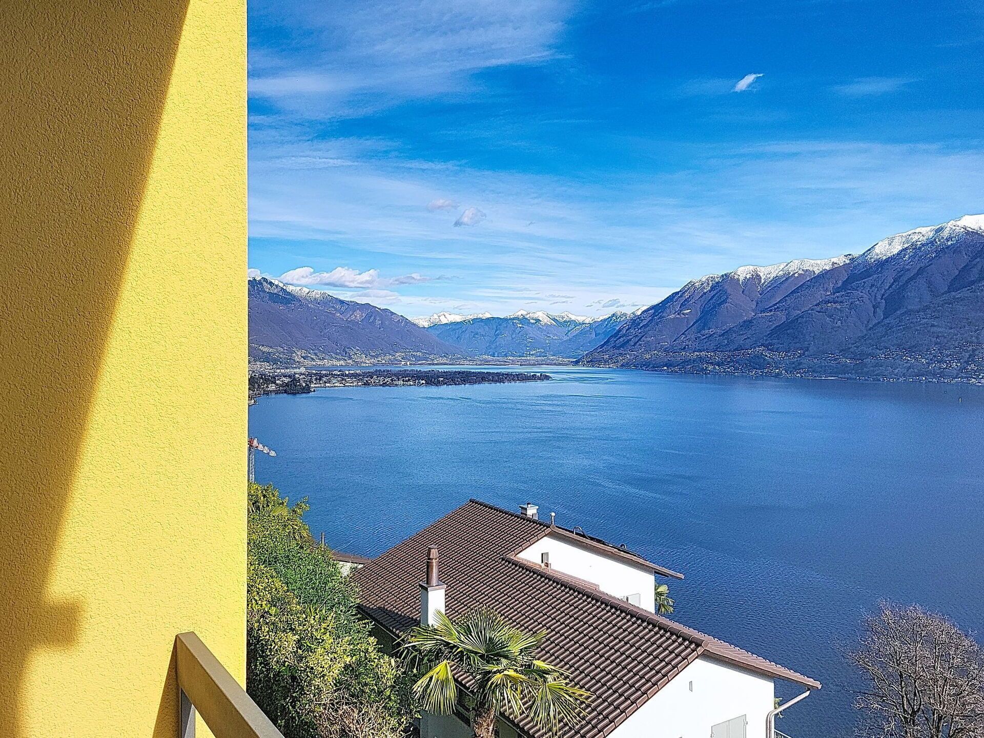 En vy över en sjö med berg i bakgrunden, en byggnad med ett tak täckt av tegelpannor och en gul vägg till vänster.