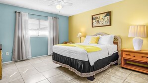 Un dormitorio con cama, ventilador de techo, ventana con cortinas, mesita de noche con lámpara y un cuadro en la pared.