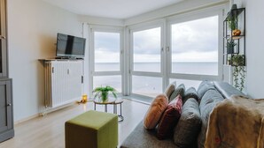 Uma sala de estar moderna com uma grande janela com vista para o mar, uma televisão de tela plana sobre um móvel, um banquinho verde e um sofá cinza com almofadas.