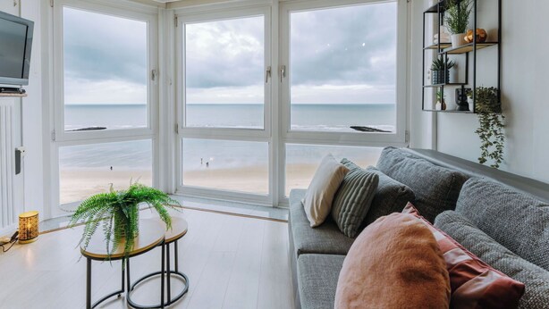 Uma sala de estar com uma grande janela com vista para a praia, um sofá cinzento, uma mesinha com uma planta e uma prateleira fixada na parede com plantas.