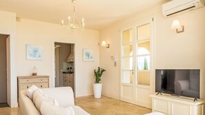 Ein Wohnzimmer mit einem weißen Sofa, einem Fernseher auf einem weißen Schrank, einer Chandelüster und einer Topfpflanze.