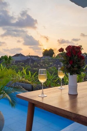 Une table en bois, sur laquelle reposent deux verres de vin blanc et un vase de roses rouges, offre une vue sur une piscine et un paysage pittoresque composé de maisons et de verdure.