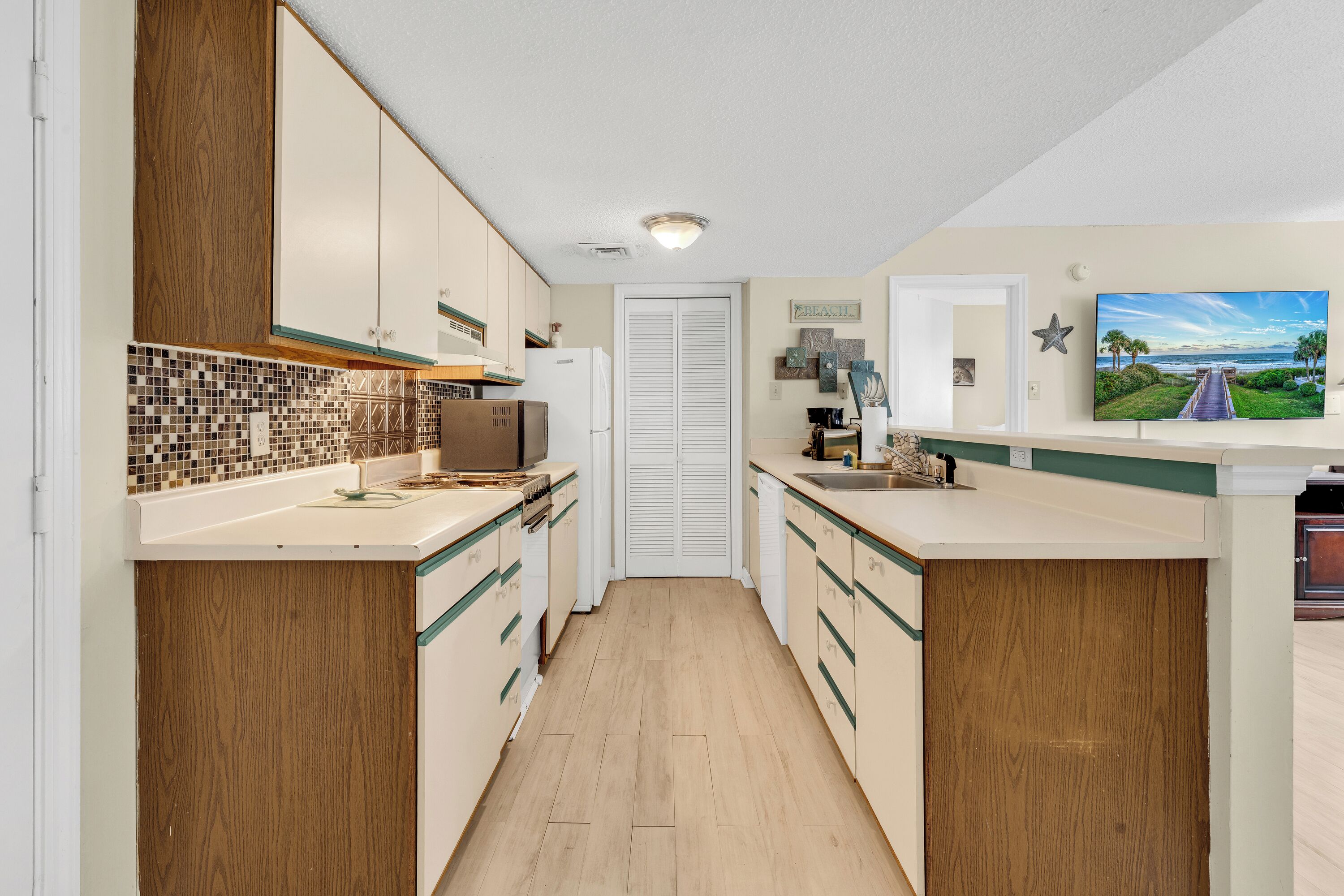 A narrow kitchen with wooden cabinets, a tiled backsplash, and a view of the beach through a window.