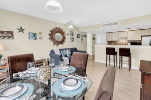 A dining area with a glass table set for four, a kitchen in the background, and a living room with a sofa and chairs. - Myrtle Beach Resort 6306 (Myrtle Beach)