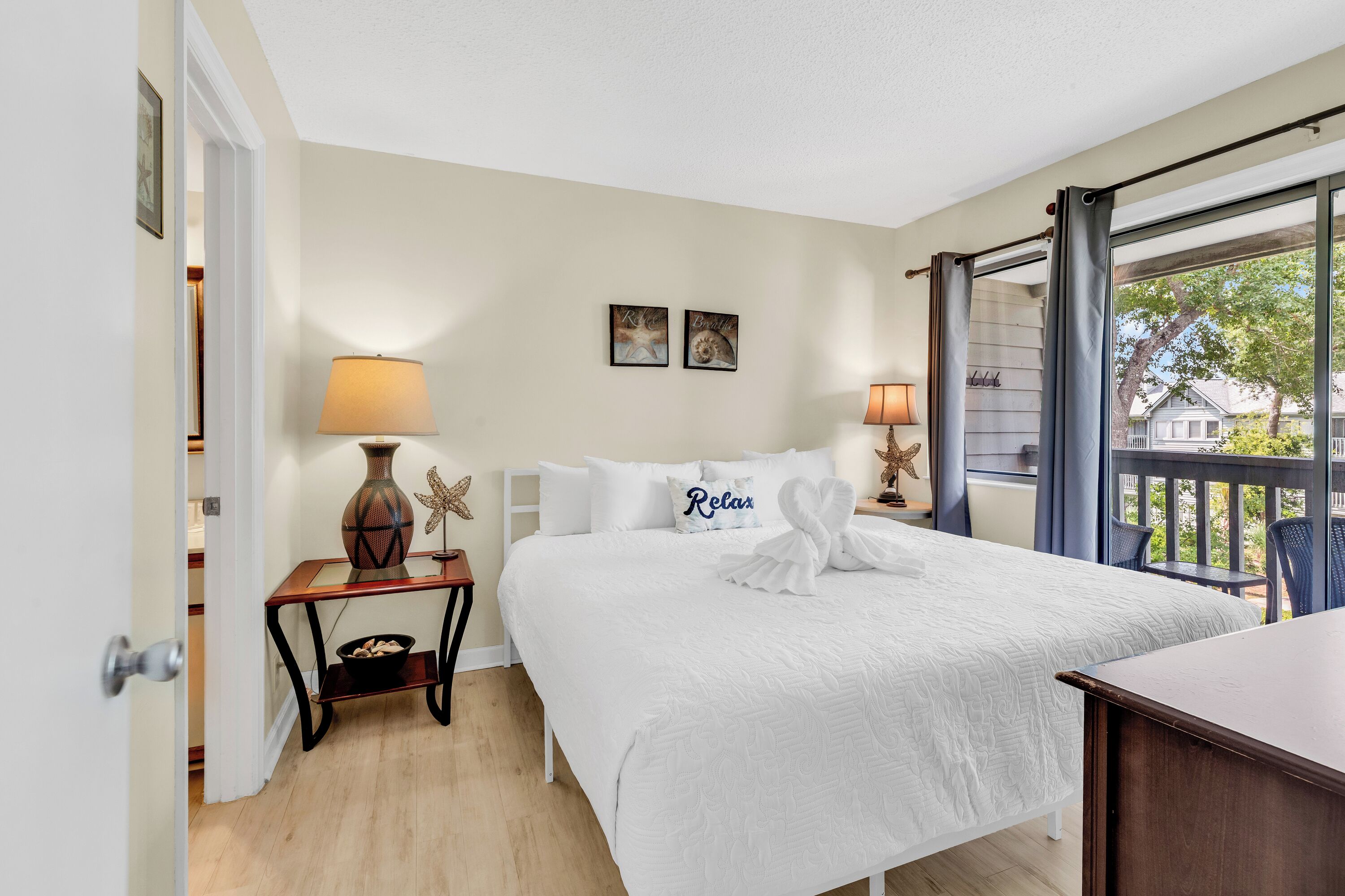 Quarto de hotel com uma cama, duas luminárias de cabeceira, uma mesa pequena com abajur, um vaso decorativo e vista para o exterior.