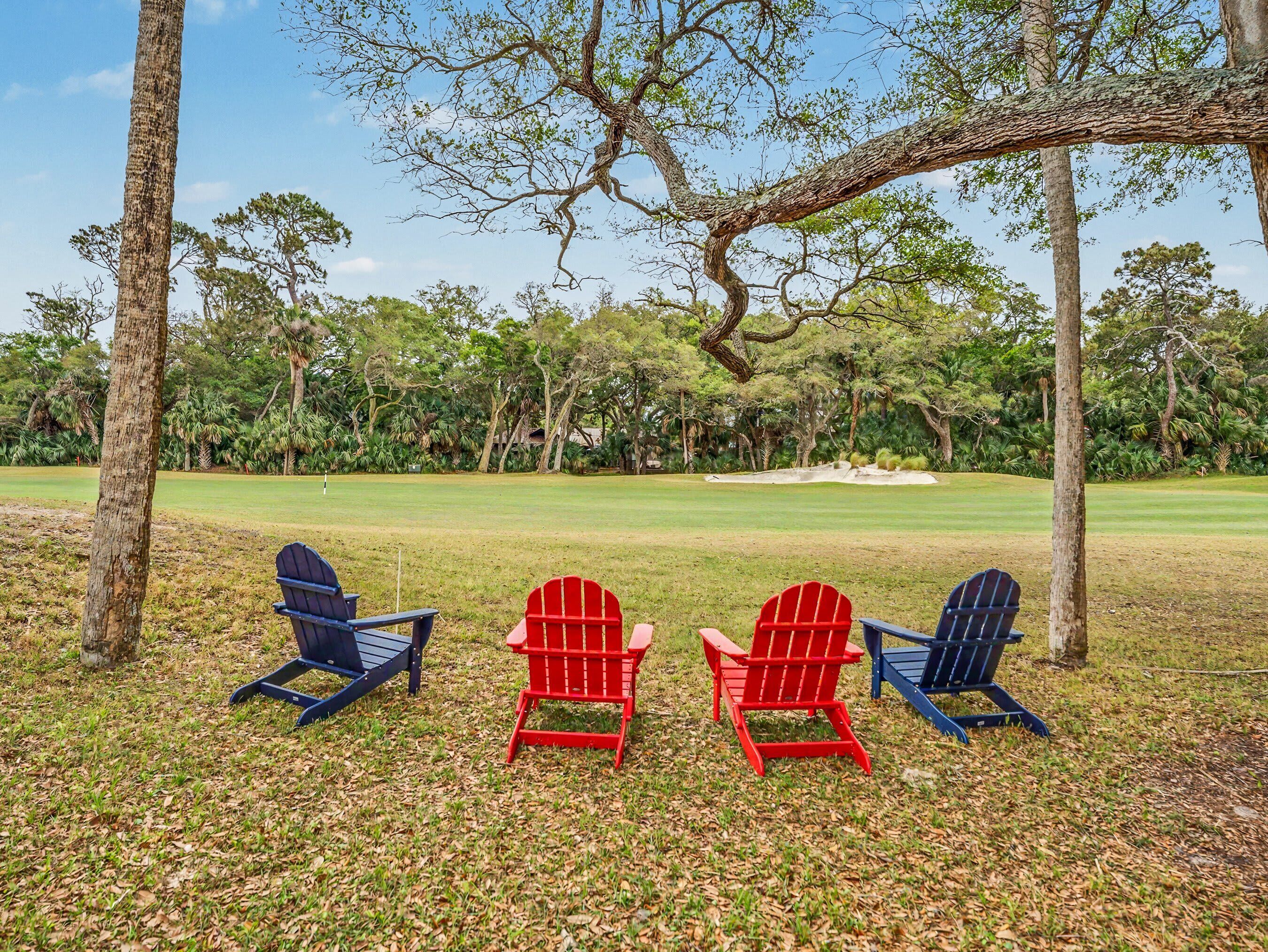 Un terrain de golf avec quatre fauteuils Adirondack, dont un rouge et trois bleus, placés sous un grand arbre.