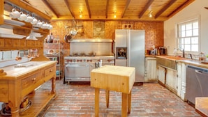 Una cocina rústica con vigas de madera, suelo de ladrillo, una estufa central y una mesa de madera.
